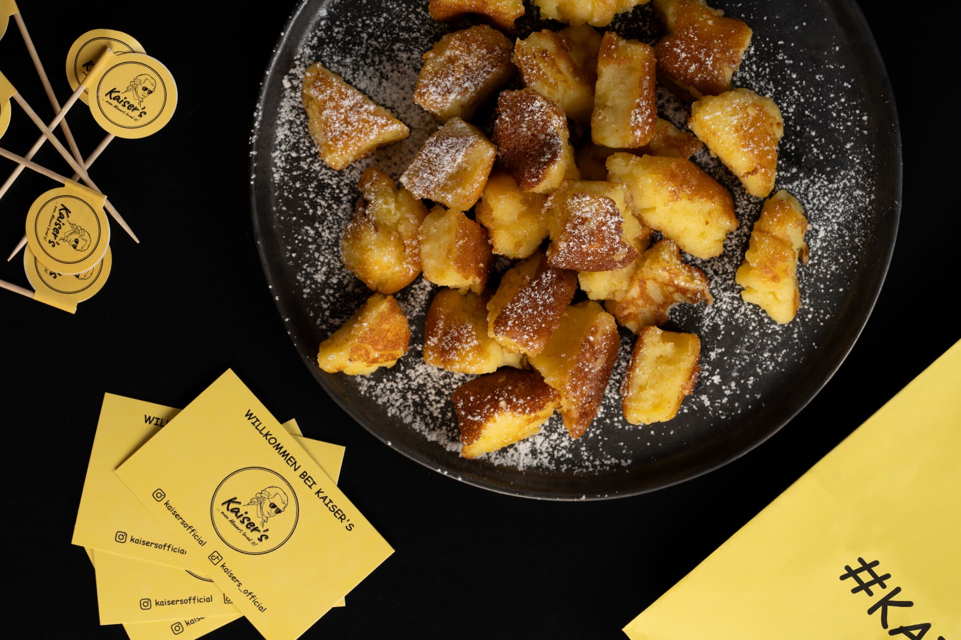 Frischer Kaiserschmarrn mit Puderzucker und gelben Kaiser’s Markenaccessoires mit Mozart-Motiv